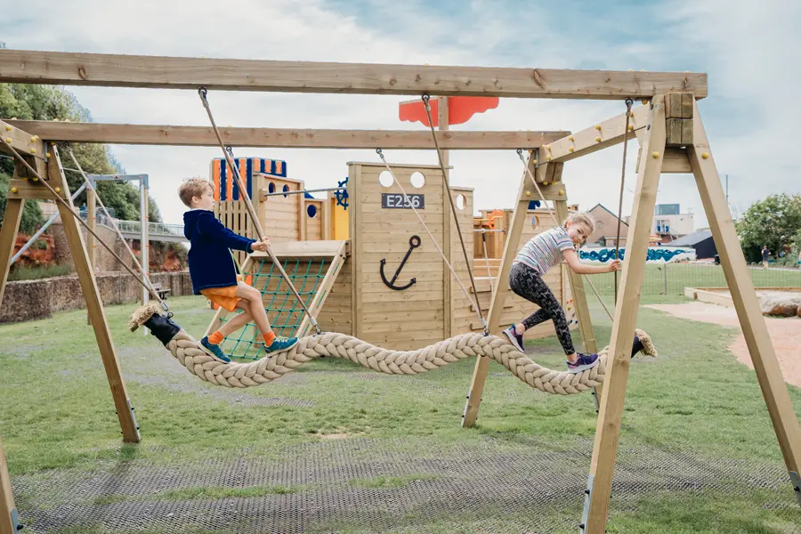 A boy and a girl balancing on a thick, braided snake rope swing, suspended from a timber frame in front of a large, ship-themed play structure in an outdoor playground.