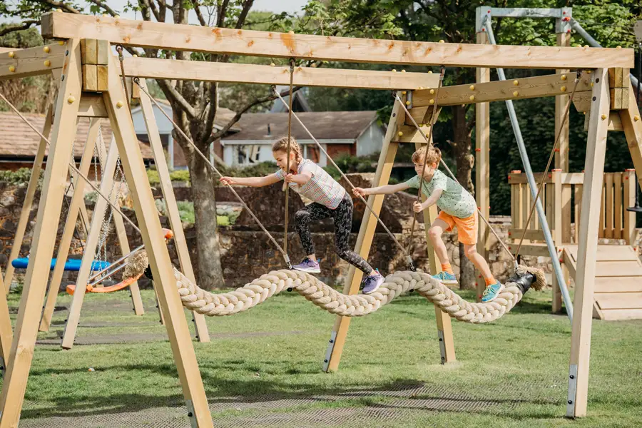 A boy and a girl holding hands while carefully walking and balancing along a thick, braided snake rope swing in a sunny, outdoor playground.