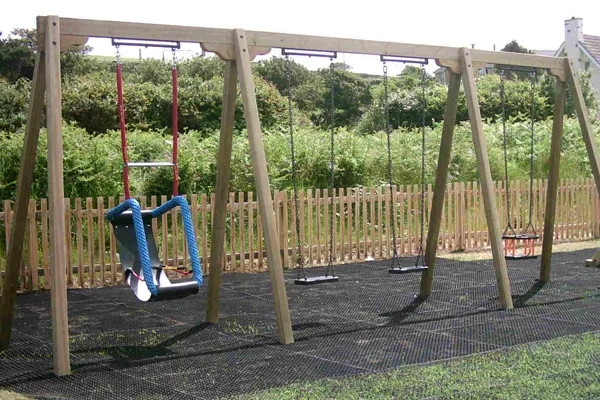 A wide view of a timber triple swing frame in a coastal playground, featuring a special needs cradle swing and two standard swings on a rubber mat.
