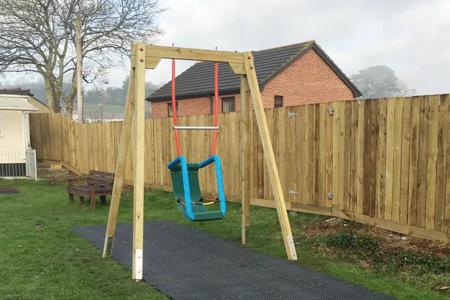 A special needs inclusive cradle swing with a green supportive seat and blue ropes, hanging from a timber A-frame on a black rubber mat in a garden playground.