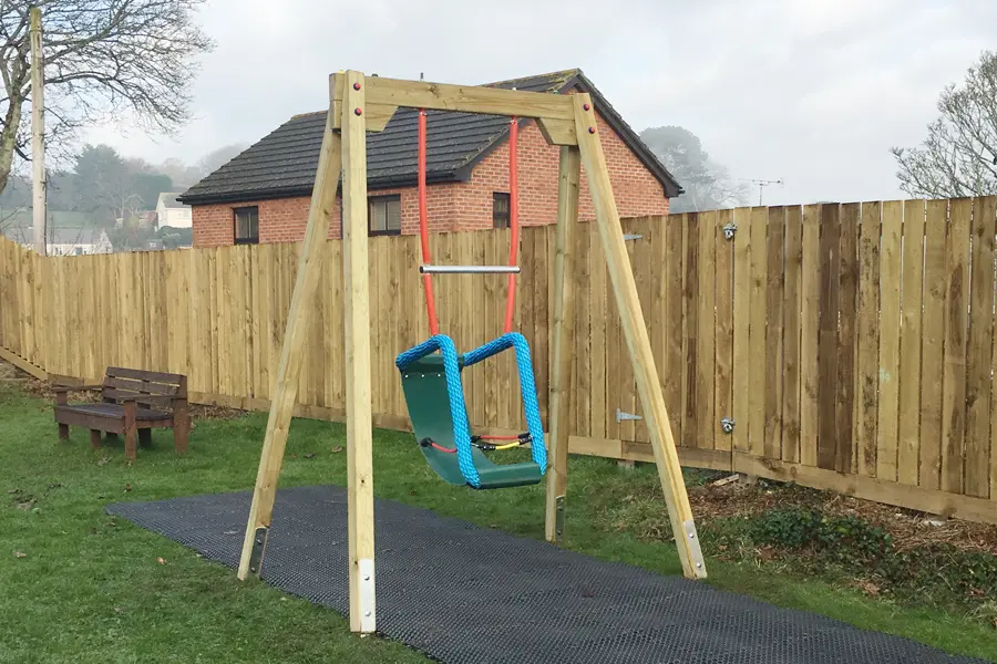 A side view of a special needs inclusive cradle swing with a green supportive seat, hanging from a timber A-frame on a rubber mat in a garden.