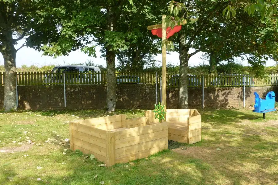 The St Columb Sailor children's outdoor play boat in a park setting, with trees and a fence in the background.