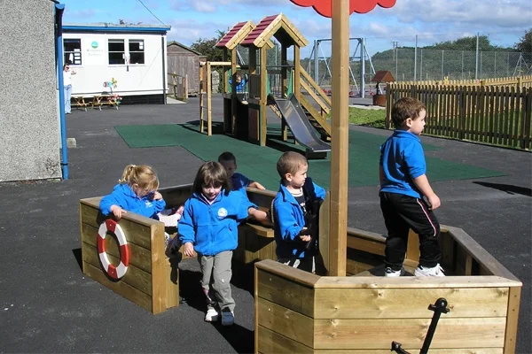 A group of school children in blue uniforms playing together in the St Columb Sailor outdoor wooden play boat.