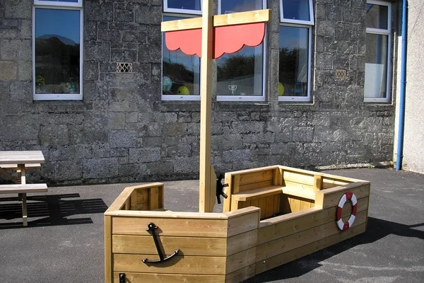 The St Columb Sailor outdoor wooden play boat in a school playground, with a stone building in the background
