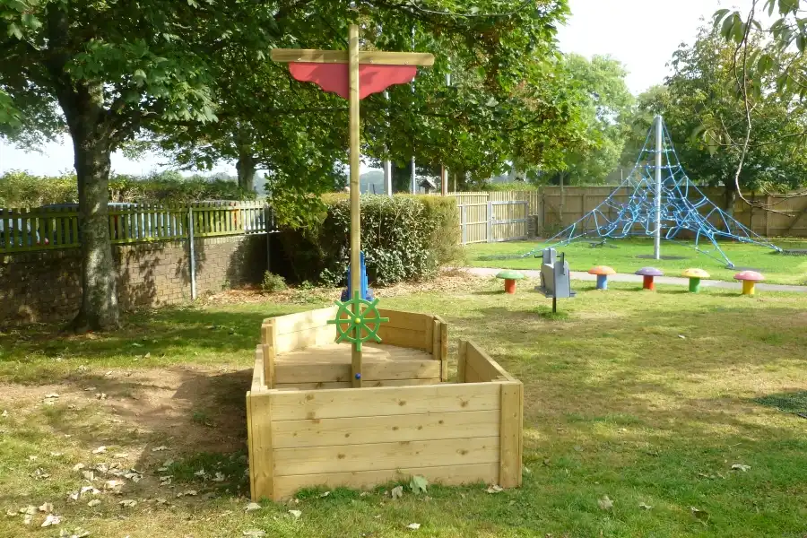 The St Columb Sailor wooden play boat and sandpit in a green outdoor playground with a climbing net in the background.