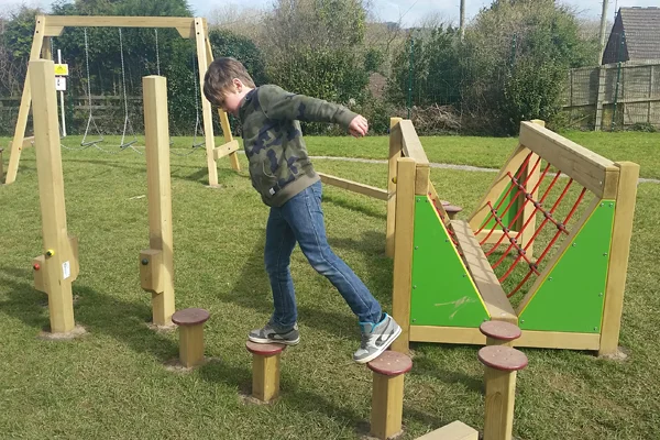 A boy stepping across a series of wooden Stepping Logs in a playground, with a V-net bridge and swings in the background.