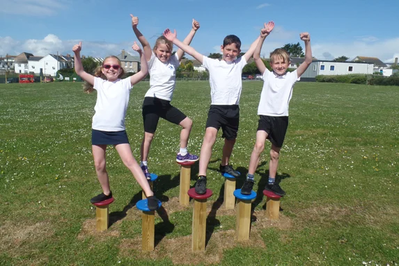 Four happy children in school uniform balancing and cheering on top of colourful Stepping Logs in a school field.