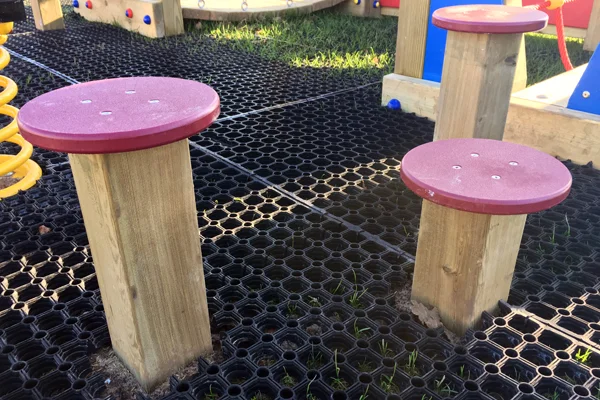 A close-up of two wooden Stepping Logs with textured red tops, installed on a black rubber grass mat safety surface.
