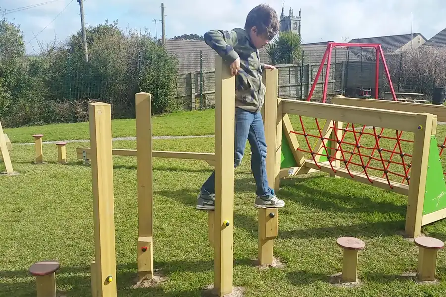 A child carefully balancing on the Stepping Stilts in a sunny outdoor playground with other trim trail items nearby.