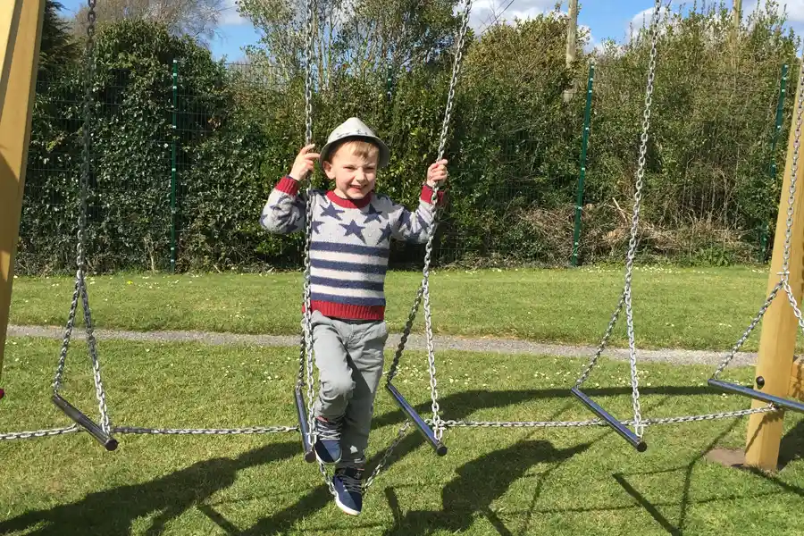 A young boy smiling as he balances on the Swinging Steps outdoor chain steps in a playground.