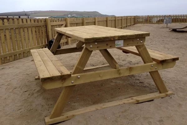 A heavy-duty timber picnic bench with an A-frame design, set on a sandy surface in a coastal playground with a timber fence in the background.