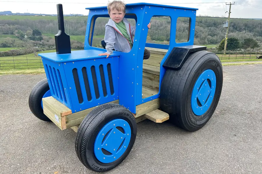 A blue timber play tractor with a child standing inside, on a tarmac surface in an outdoor playground.