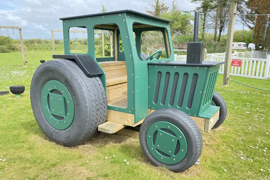 A green and black timber play tractor with an open cab, on a grassy area in an outdoor playground.