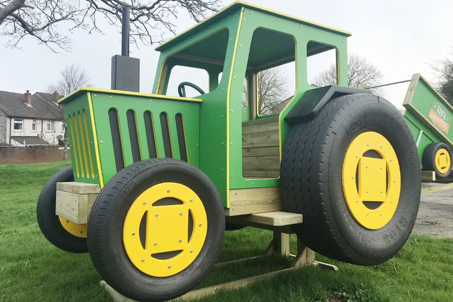 A green and yellow timber play tractor with a tipper trailer, on a grassy area in an outdoor playground.
