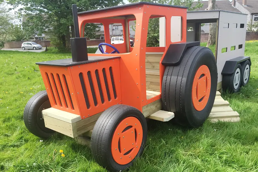 An orange and black timber play tractor with a grey livestock trailer, on a grassy area in an outdoor playground.