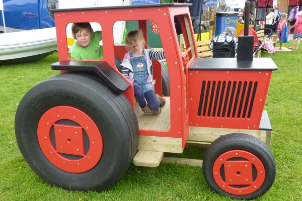 A red and black timber play tractor with two children inside, on a grassy area in an outdoor playground.