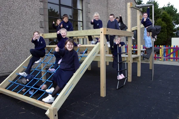 A group of uniformed school children happily playing on the Torpoint Activity Centre timber climbing frame, climbing the rope net and hanging from the bars in their school playground.