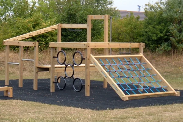 The Torpoint Activity Centre timber climbing frame from another angle, showing the blue rope net, monkey rings, and overhead ladders, on a black rubber safety surface in a school playground.