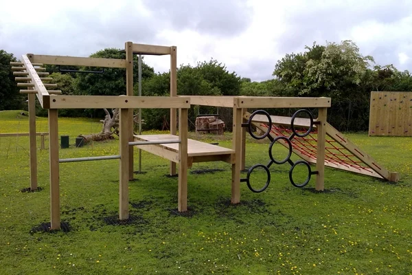 The Torpoint Activity Centre, a timber climbing frame with monkey rings, a red rope net, a climbing wall, and overhead ladders, set on rubber mulch in a grassy outdoor playground.
