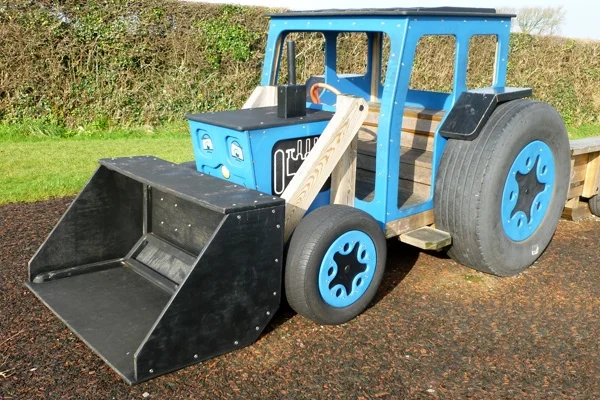 A child climbing on a timber play tractor with a front bucket loader, on a rubber surface in an outdoor playground.
