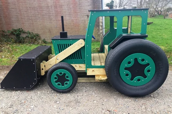 A side view of a timber play tractor with a front bucket loader, on a grassy area in an outdoor playground.