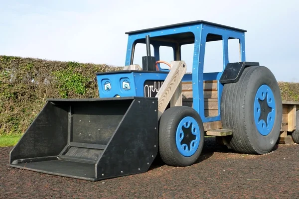 A rear view of a timber play tractor with a front bucket loader, on a grassy area in an outdoor playground.