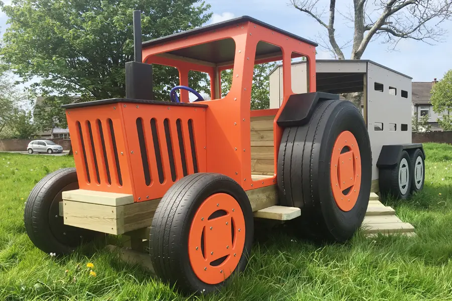 An orange and black timber play tractor with a grey livestock trailer slide, on a grassy area in an outdoor playground.