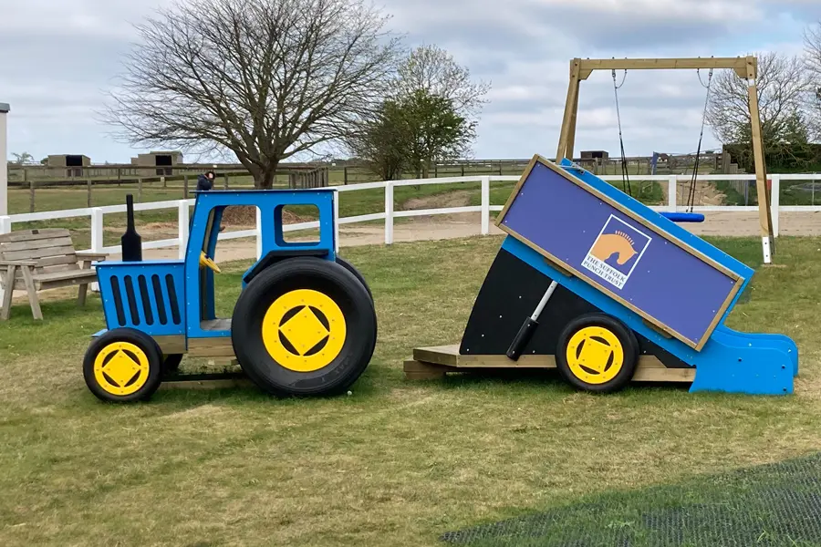 A blue and yellow timber play tractor with a tipper trailer slide, on a grassy area in an outdoor playground.