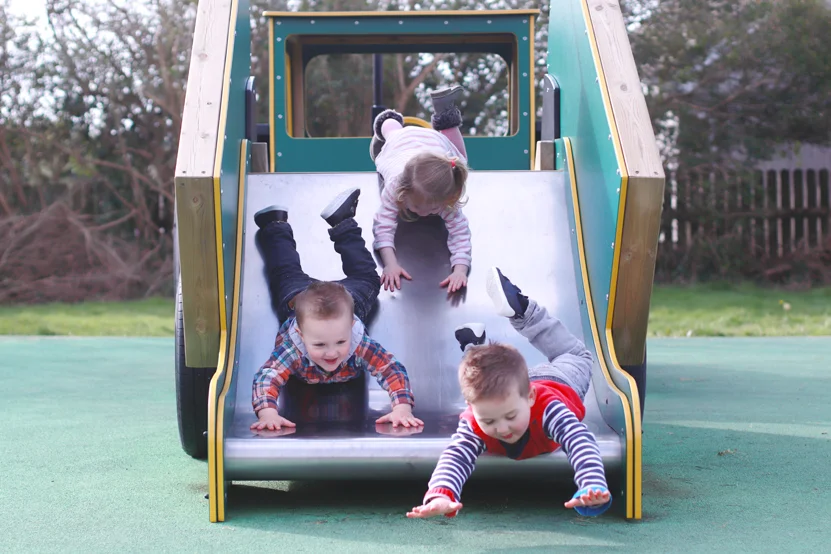 Three young children sliding down a green and yellow timber tractor trailer slide in an outdoor playground.