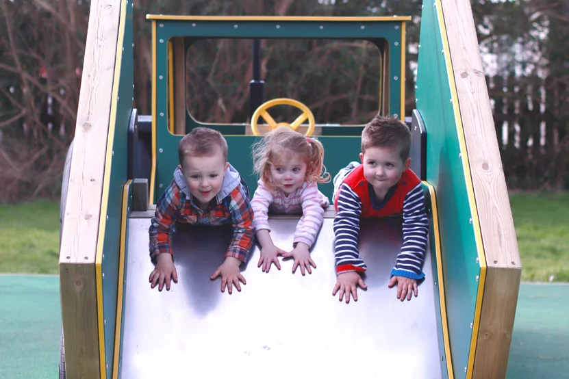 Three young children at the top of a green and yellow timber tractor trailer slide in an outdoor playground.