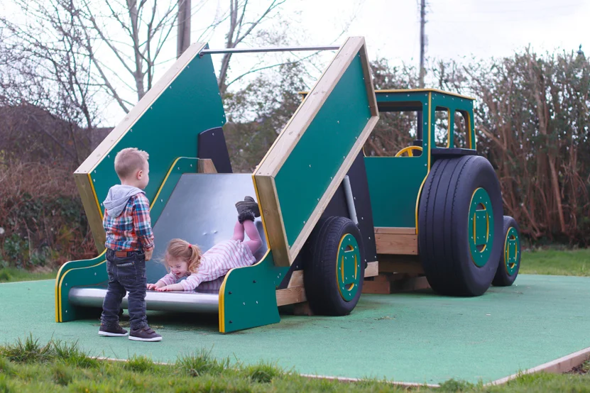 A green and yellow timber tractor and tipper trailer slide, with two children playing on it in an outdoor playground.