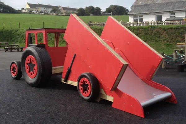 A red and black timber tractor with a tipper trailer slide, on a tarmac surface in an outdoor playground.