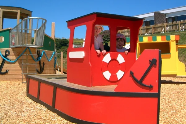 Two happy children playing in a bright red Trawler Sandpit outdoor play boat at a playground.