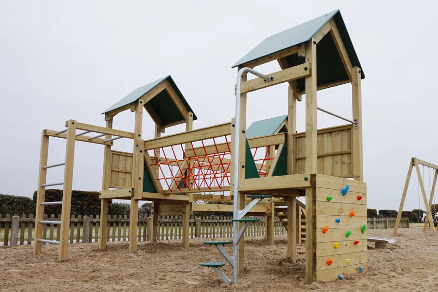 A close-up side view of the Trevean Towers showing the red rope climbing net, colourful rock climbing wall, galvanised monkey bars, and green-roofed towers, with a swing set visible in the background.