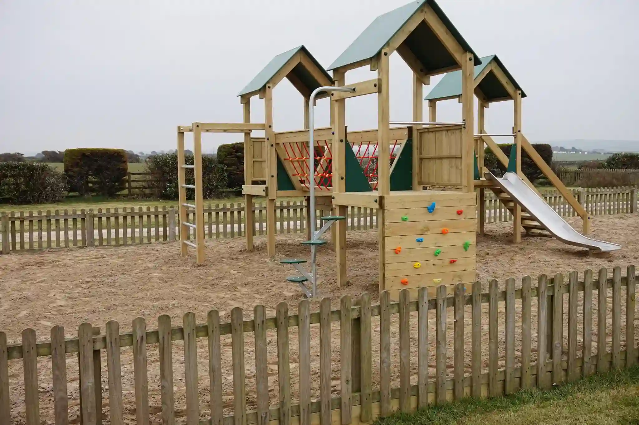 A wide-angle view of the Trevean Towers multi-play unit showing the colourful climbing wall, stainless steel slide, red rope climbing net, and three green-roofed towers, surrounded by a timber picket fence with Cornish countryside in the background.