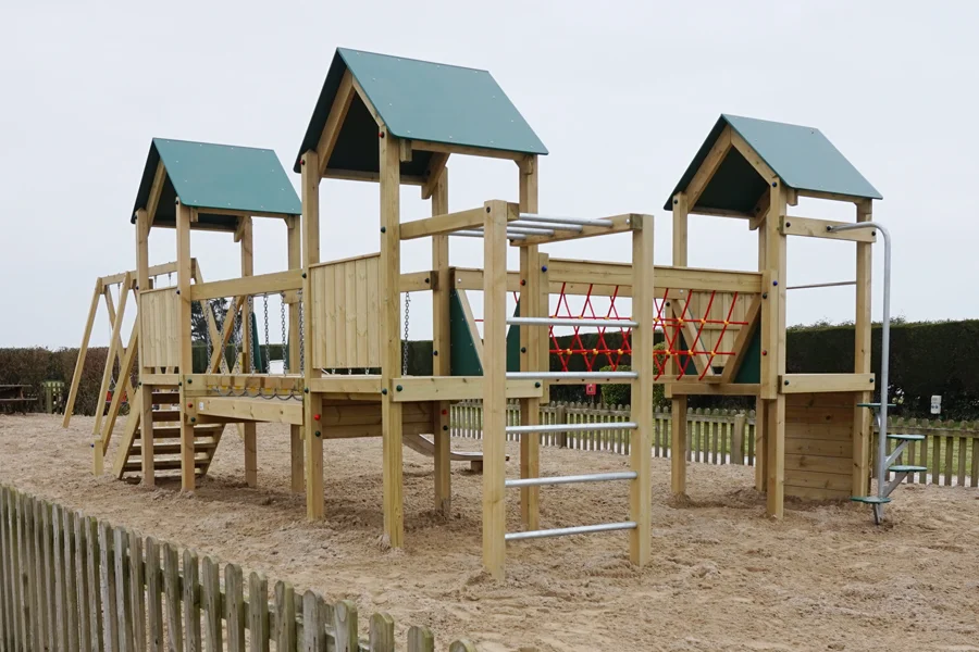A rear view of the Trevean Towers multi-play unit showing the chain bridge, red rope climbing net, monkey bars, and three green-roofed towers, in a sand playground surrounded by a timber picket fence.