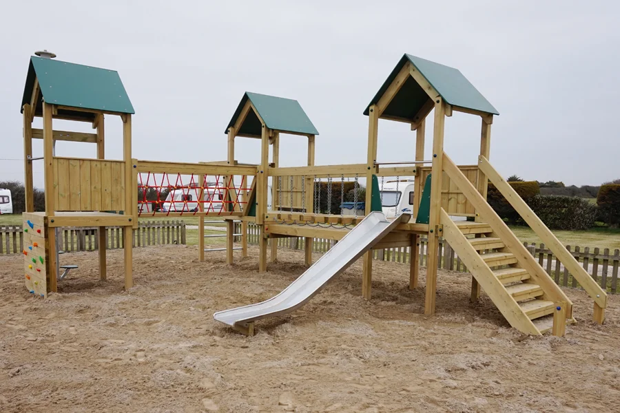 A large wooden Trevean Towers multi-play unit with three green-roofed towers, a stainless steel slide, climbing wall, and red rope net, installed in a sand-surfaced playground at a caravan park.