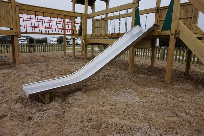 A close-up ground-level view of the stainless steel slide on the Trevean Towers multi-play unit, with the red rope climbing net and chain swings visible in the background, in a sand playground at a caravan park.