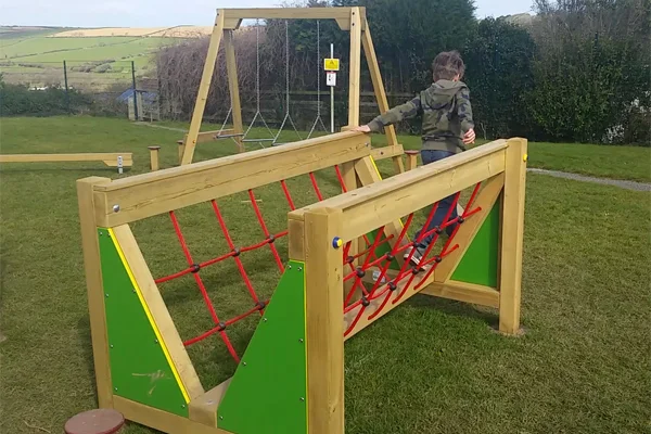 A young boy in a camouflage hoodie carefully walks across the V Net Bridge Low Level in a park.