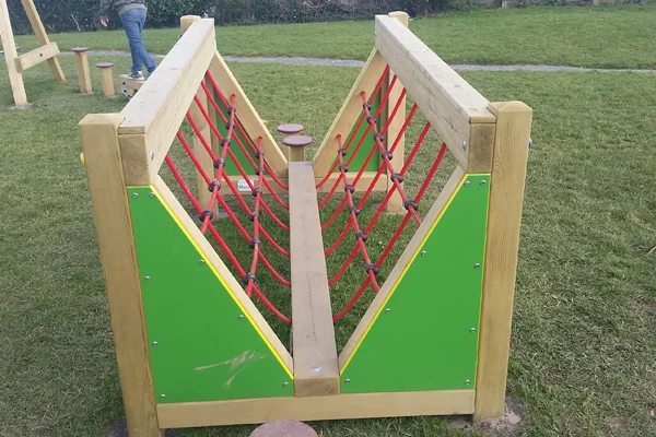 A close-up, end-on view of the V Net Bridge Low Level, showing the wooden balance beam and red rope safety netting.
