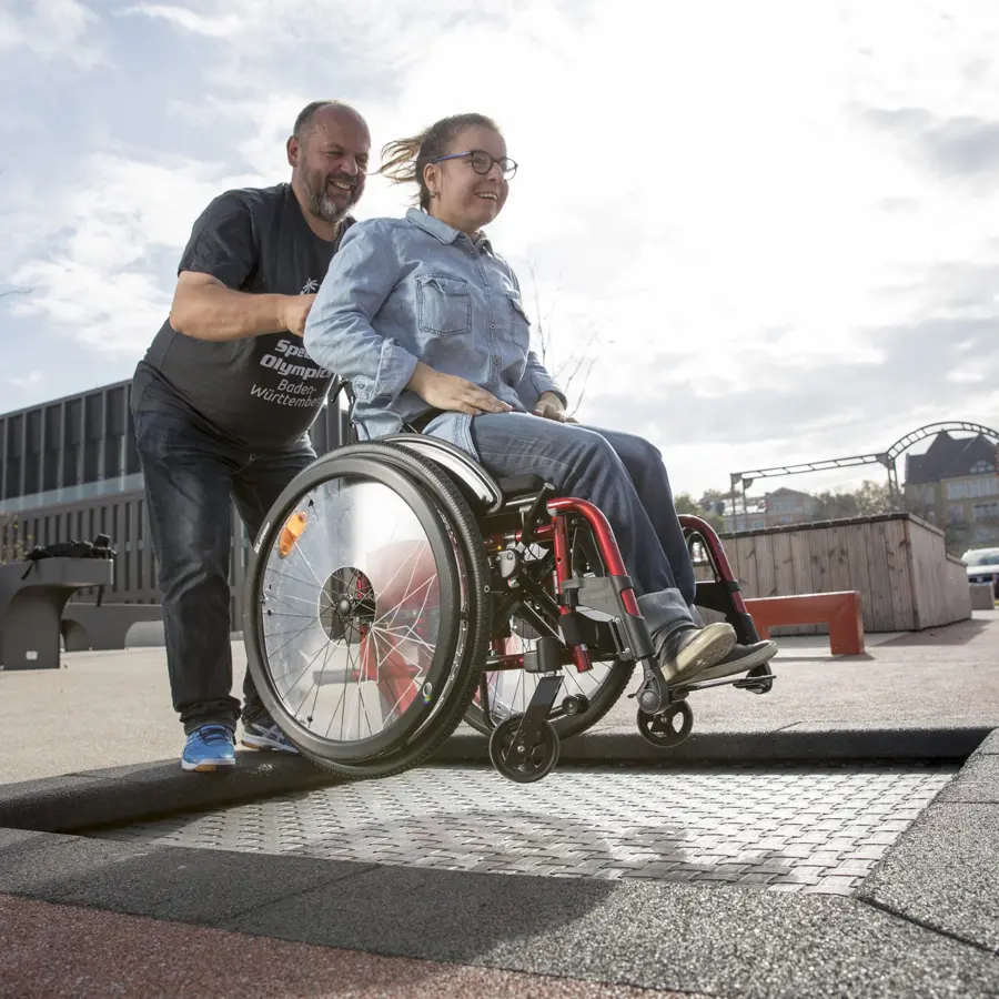 A woman in a wheelchair smiling as she bounces on a flush-mounted, inclusive wheelchair trampoline, assisted by a man in an outdoor playground setting.