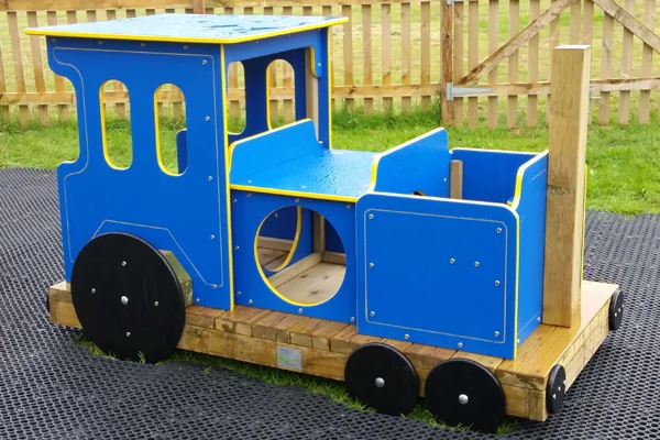 Blue wooden children's play train with black wheels on outdoor rubber matting near a wooden fence