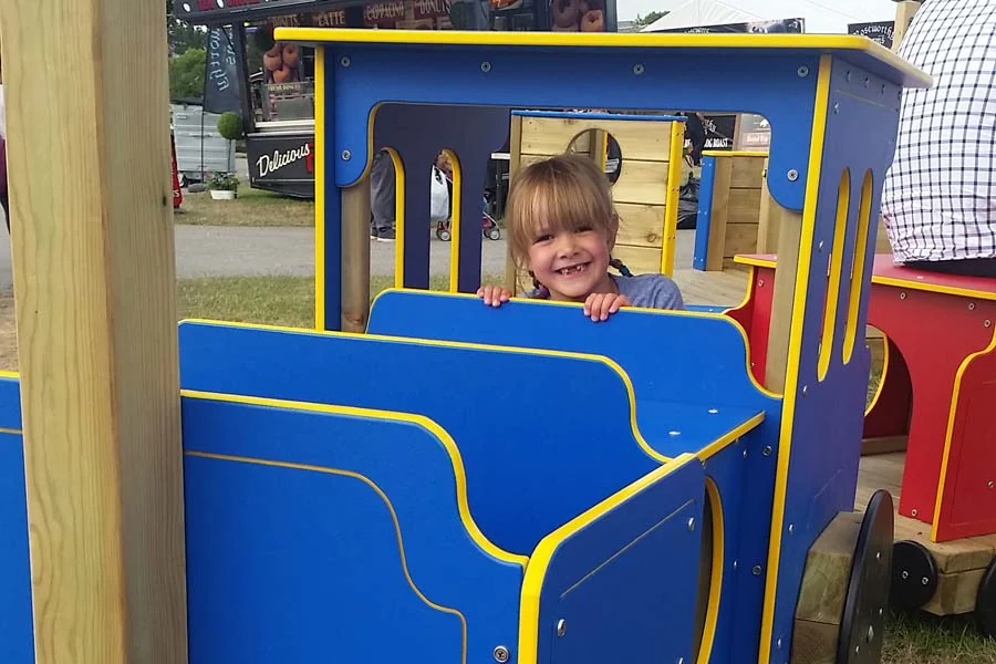 Smiling child peeking over the side of a colorful blue and yellow miniature train car at an outdoor park.
