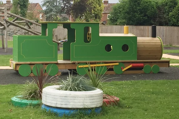 Green and yellow wooden miniature steam train on a track in a grassy park with playground equipment and houses in the background