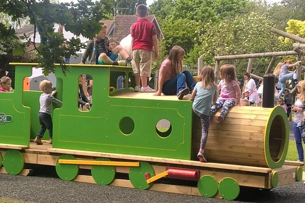 Children playing on a green and wooden miniature steam train in a park playground with trees and climbing frames in the background