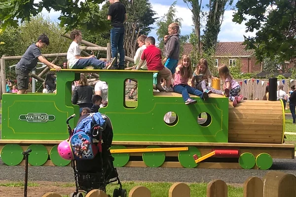 Children playing on a green and wooden miniature train in a sunny park with trees and houses in the background.