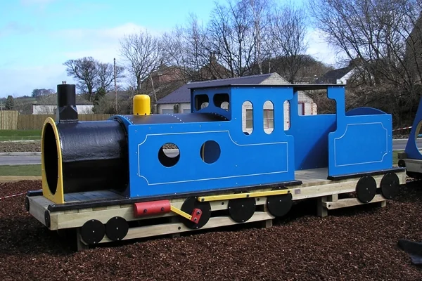 Blue and black miniature steam train engine on wooden track in outdoor park setting with bare trees and houses in background