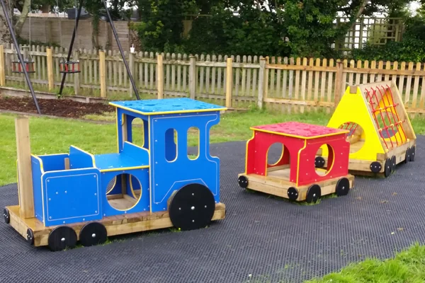 Colorful wooden miniature train set on rubber matting in a grassy outdoor playground with swings and wooden fence in background