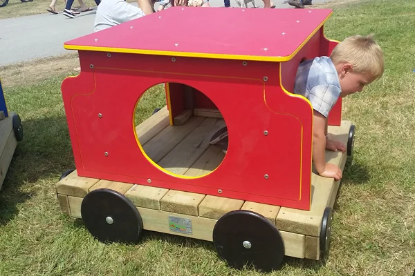 Child playing inside a red and yellow wooden toy train on grass in a park setting.