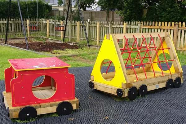 Colorful wooden playground train with red and yellow sections on a rubber mat in a fenced grassy area